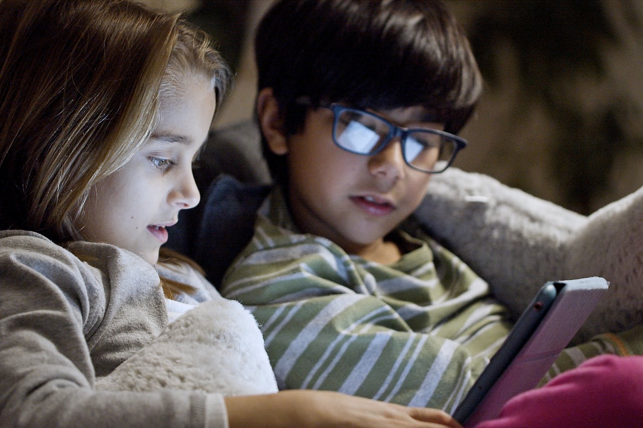 Two children comfortably using a tablet in bed at night in a cozy setting.